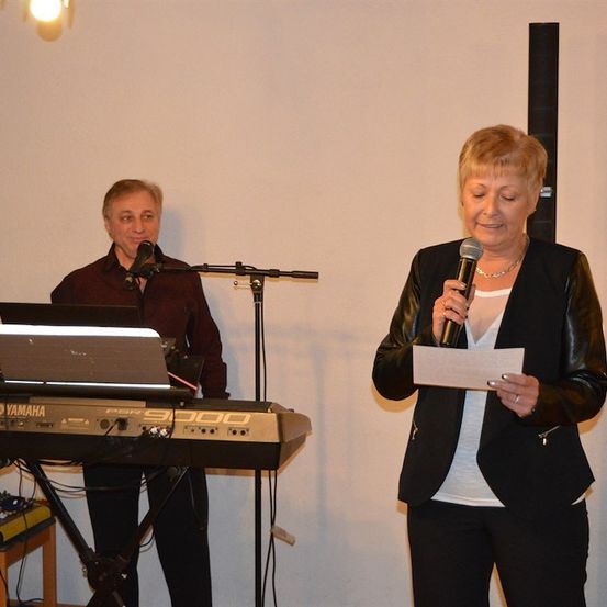 Ein Mann und eine Frau stehen vor einem Yamaha-Keyboard. Der Mann spielt das Keyboard, während die Frau ein Mikrofon und ein Stück Papier in der Hand hält.