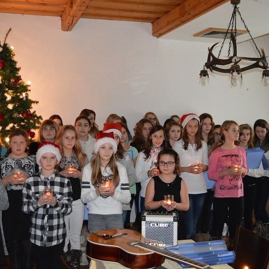 Eine Gruppe von Kindern in Weihnachtsmützen und mit Kerzen steht in einem Raum mit einem Weihnachtsbaum und einer Gitarre auf einem Tisch.