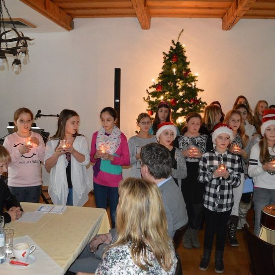 Eine Gruppe von Kindern und Erwachsenen versammelt sich um einen Weihnachtsbaum, hält Kerzen und singt. Der Raum ist für Weihnachten geschmückt, mit einem Tisch und Stühlen im Vordergrund.