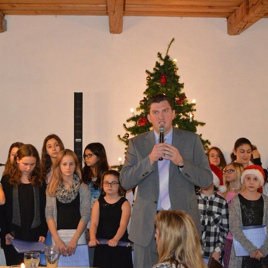 Ein Mann in einem Anzug spricht in ein Mikrofon vor einem Weihnachtsbaum mit einer Gruppe von Kindern und Erwachsenen.
