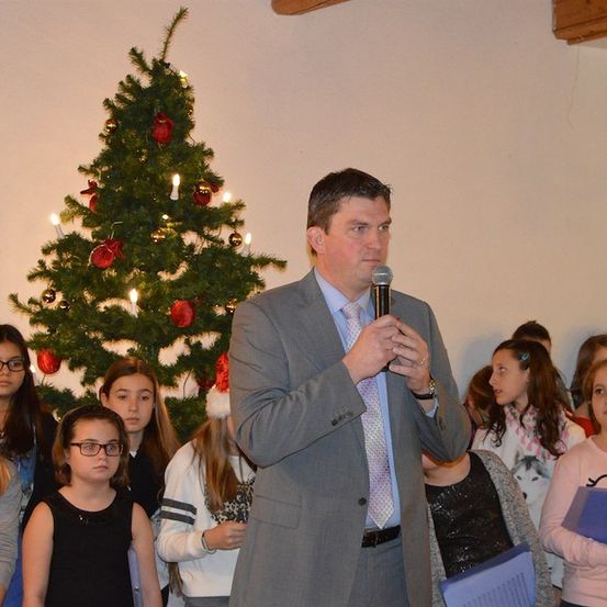 Ein Mann in einem Anzug spricht in ein Mikrofon vor einer Weihnachtsbaum mit Kindern, die zuhören.