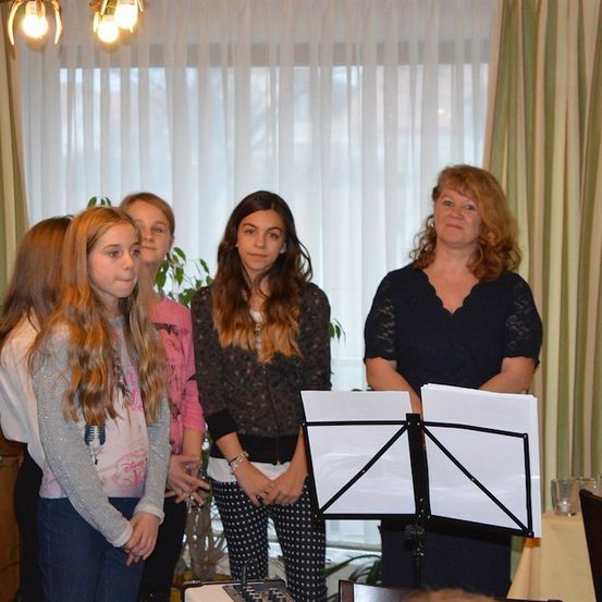 Eine Gruppe junger Mädchen und eine Frau stehen vor einem Notenständer mit Papieren darauf. Sie sind in einem Raum mit einem Fenster und Vorhängen.