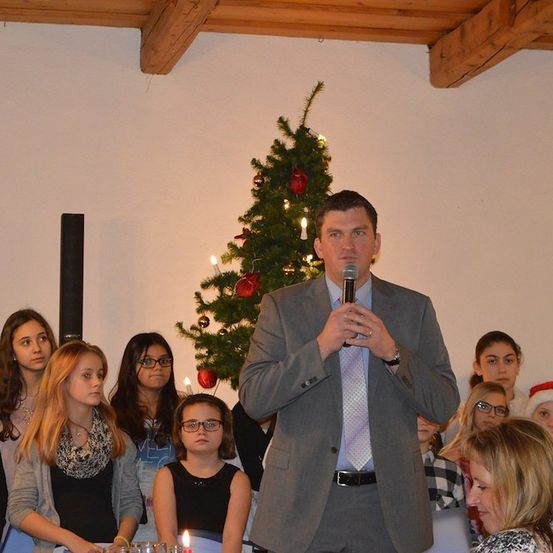 Ein Mann in einem Anzug spricht in ein Mikrofon vor einem geschmückten Weihnachtsbaum, während mehrere junge Mädchen aufmerksam zuhören.