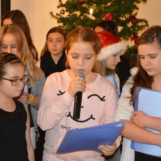 Eine Gruppe junger Mädchen versammelt sich um einen Weihnachtsbaum, ein Mädchen hält ein Mikrofon und singt, während andere blaue Ordner halten und zuhören.