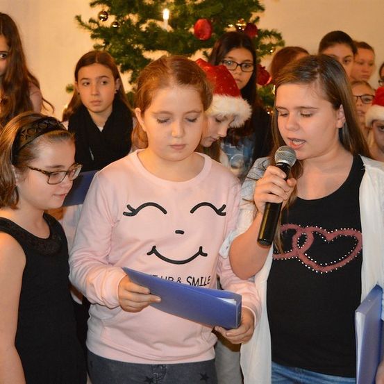 Eine Gruppe junger Mädchen singt Weihnachtslieder in einem Raum mit einem Weihnachtsbaum im Hintergrund. Eines der Mädchen hält ein Mikrofon und ein anderes liest von einem Stück Papier.