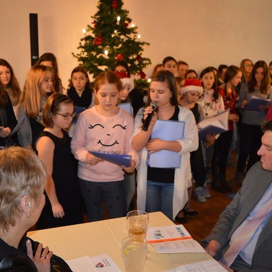 Eine Gruppe von Kindern und Erwachsenen versammelt sich um einen Weihnachtsbaum, singt und hält Papiere in der Hand. Ein Mann in Anzug sitzt an einem Tisch mit einem Glas Wasser und Papieren.