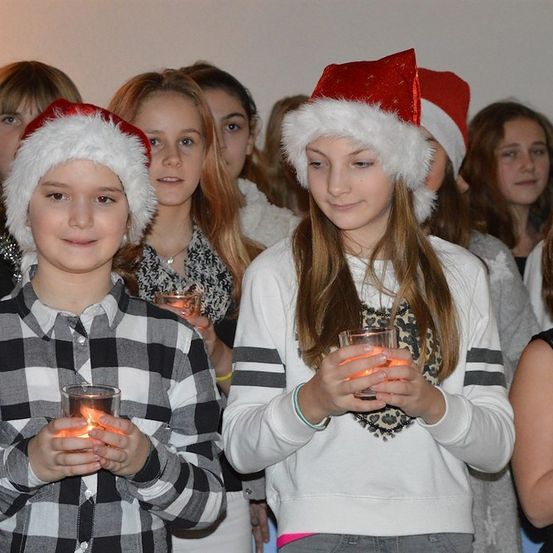 Eine Gruppe von Kindern mit Weihnachtsmützen hält Kerzen, möglicherweise bei einer Weihnachtsveranstaltung. Das Mädchen auf der rechten Seite lächelt, während es eine Kerze hält.