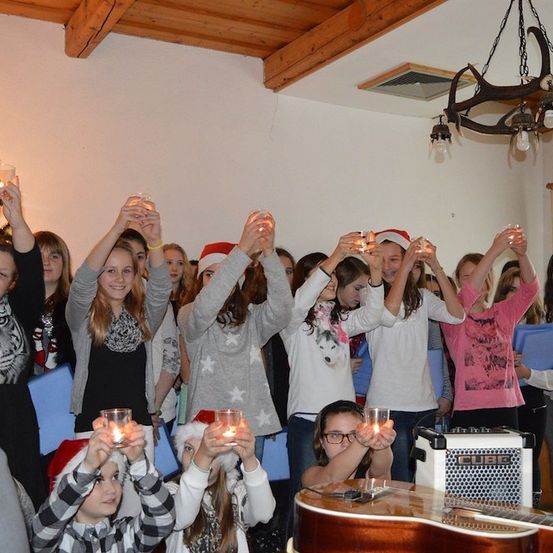 Eine Gruppe von Kindern und Jugendlichen versammelt sich drinnen, hält brennende Kerzen in den Händen, ist festlich gekleidet und singt Weihnachtslieder. Ein Weihnachtsbaum und eine Gitarre sind im Hintergrund zu sehen.