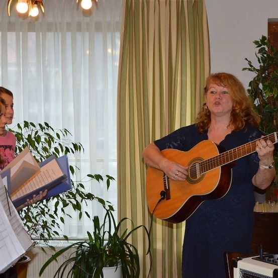 Eine Frau spielt Gitarre und singt, während einige Leute zusehen und Noten halten.