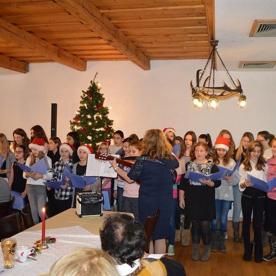 Eine Gruppe von Kindern und Erwachsenen singt Weihnachtslieder in einem Raum mit einem Weihnachtsbaum und einer Kronleuchter. Einige halten Notenblätter.