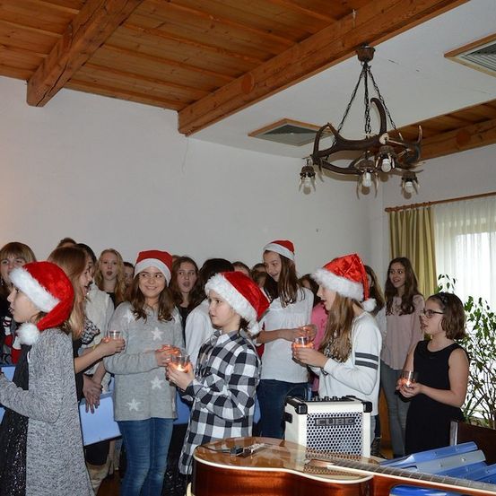 Eine Gruppe junger Mädchen mit Weihnachtsmützen versammelt sich in einem Raum, hält Kerzen und singt Weihnachtslieder. Ein Weihnachtsbaum und eine Geweih-Kronleuchter sind im Hintergrund.