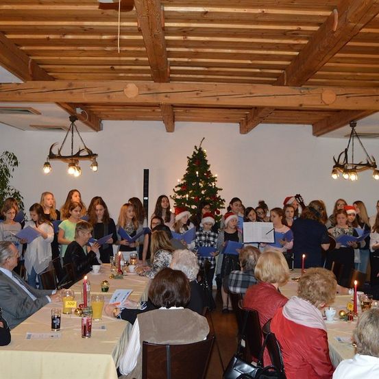 Eine Gruppe von Menschen versammelt sich in einem Raum und singt Weihnachtslieder. Sie halten Bücher in den Händen und stehen um einen Weihnachtsbaum herum. Der Raum ist mit Weihnachtslichtern und Kerzen geschmückt.