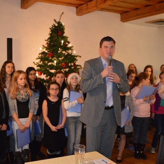 Ein Mann in einem Anzug steht vor einem Mikrofon und spricht zu einer Gruppe junger Mädchen, die blaue Ordner halten. Ein Weihnachtsbaum steht dahinter.