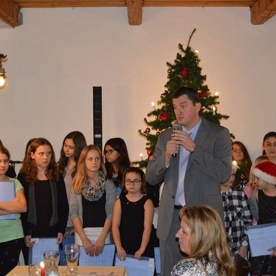 Ein Mann in einem Anzug spricht in ein Mikrofon vor einer Gruppe von Kindern und Erwachsenen, mit einem Weihnachtsbaum und Dekorationen im Hintergrund.