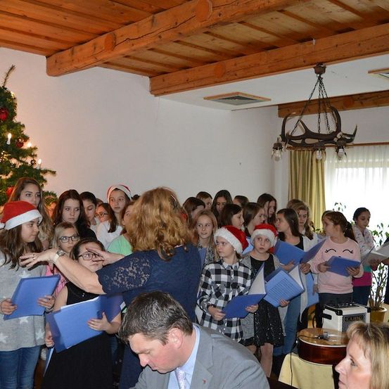Eine Gruppe von Kindern und Erwachsenen in einem Raum mit einem Weihnachtsbaum, die Weihnachtsmützen tragen, blaue Liederbücher halten und singen. Eine Frau in einem blauen Kleid führt sie an. Ein Mann in einem Anzug sitzt vor ihnen. Ein Weihnachtsbaum ist mit Lichtern und Ornamenten geschmückt.
