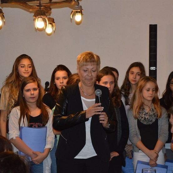 Eine Frau steht vor einer Gruppe junger Mädchen, hält ein Mikrofon in der Hand und spricht. Die Mädchen stehen in einer Reihe und hören aufmerksam zu. Der Raum ist gut beleuchtet und hat eine Kronleuchter, die von der Decke hängt.