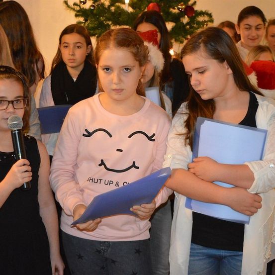 Eine Gruppe junger Mädchen steht zusammen, hält blaue Papiere in der Hand und singt vor einem Weihnachtsbaum.