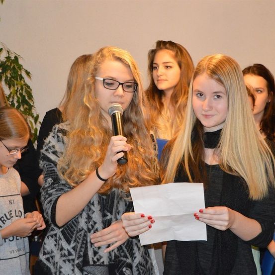 Zwei junge Frauen stehen vor einer Gruppe, eine hält ein Mikrofon und die andere ein Stück Papier. Sie scheinen eine Veranstaltung zu präsentieren oder daran teilzunehmen.