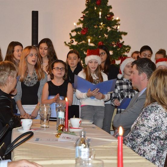 Ein Chor junger Mädchen in Weihnachtsmützen singt um einen geschmückten Weihnachtsbaum, mit Erwachsenen in der Nähe. Die Umgebung umfasst einen Tisch mit Kerzen, Tassen und Papieren.