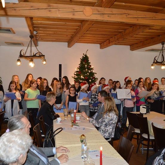 Ein Chor junger Mädchen in Weihnachtskleidung tritt in einem festlich geschmückten Raum mit einem Weihnachtsbaum und Kerzen auf den Tischen auf.