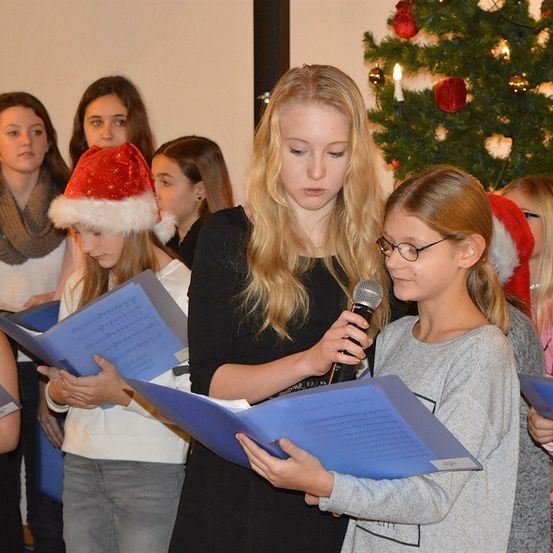 Eine Gruppe junger Mädchen singt Weihnachtslieder, während sie blaue Liederbücher halten und vor einem geschmückten Weihnachtsbaum stehen.