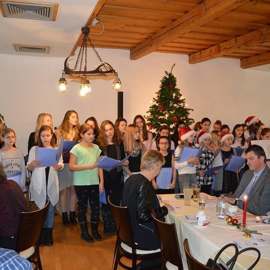 Eine Gruppe junger Mädchen und Jungen singt Weihnachtslieder in einem Raum mit einem Weihnachtsbaum und einem Mann und einer Frau, die an einem Tisch sitzen.