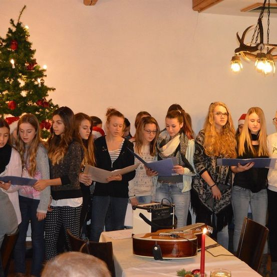 Eine Gruppe junger Frauen steht um einen Tisch herum, hält Notenblätter in den Händen und singt gemeinsam in einem für Weihnachten geschmückten Raum.