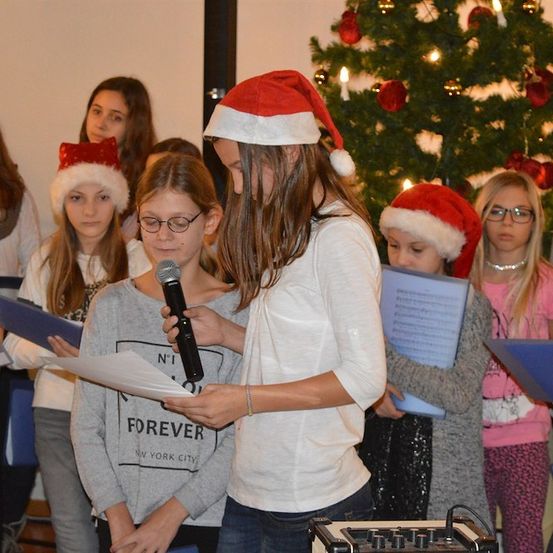 Eine Gruppe junger Mädchen mit Weihnachtsmützen singt Weihnachtslieder vor einem geschmückten Weihnachtsbaum.