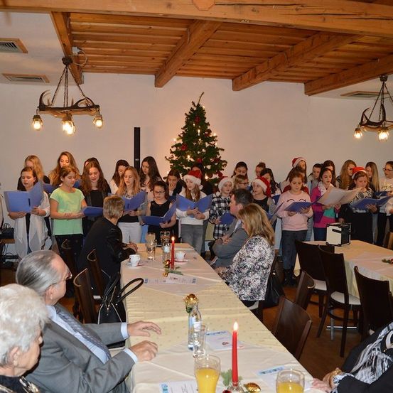 Eine Gruppe von Menschen, einschließlich Kindern und Erwachsenen, versammelt sich in einem Raum für eine Weihnachtsfeier. Sie halten blaue Liederbücher und singen, während sie vor einem geschmückten Weihnachtsbaum stehen. Der Raum hat Holzbalken und Kronleuchter.