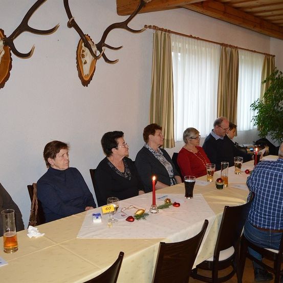 Eine Gruppe von Menschen sitzt um einen Tisch in einem Raum mit an der Wand montierten Hirschgeweihen. Es gibt Kerzen, Gläser und die Nummer 60 auf dem Tisch.