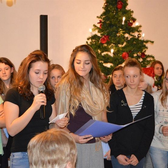 Eine Gruppe junger Frauen steht vor einem geschmückten Weihnachtsbaum, eine hält ein Mikrofon und eine andere ein Stück Papier. Sie scheinen sich auf eine Aufführung vorzubereiten.