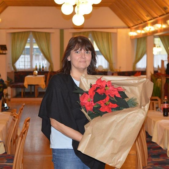 Eine Frau in einer schwarzen Jacke hält einen Strauß roter Blumen in einem Restaurant. Das Restaurant hat Holzböden, Stühle und Tische. Es gibt Flaschen und eine Lampe auf den Tischen.