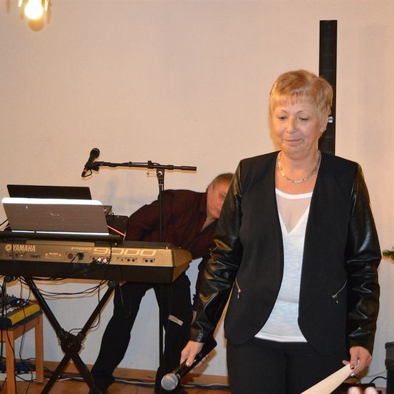 Eine Frau mit einem Mikrofon steht vor einem Yamaha-Keyboard, während ein Mann das Keyboard hinter ihr justiert. Rechts steht ein Weihnachtsbaum.