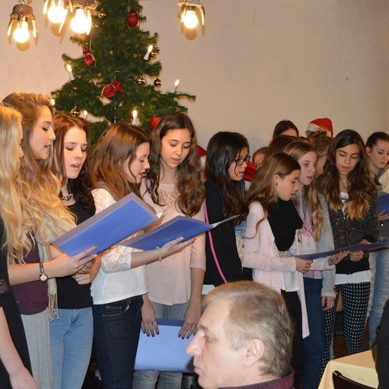 Eine Gruppe junger Mädchen in Winterkleidung singt Weihnachtslieder in einem Raum mit einem Weihnachtsbaum im Hintergrund.