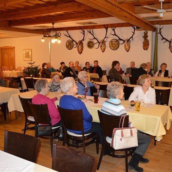 Eine Gruppe älterer Frauen und Männer sitzt an Tischen in einem Speisesaal, einige unterhalten sich und andere essen. Der Raum hat Holzbalken und Geweihe an der Wand.