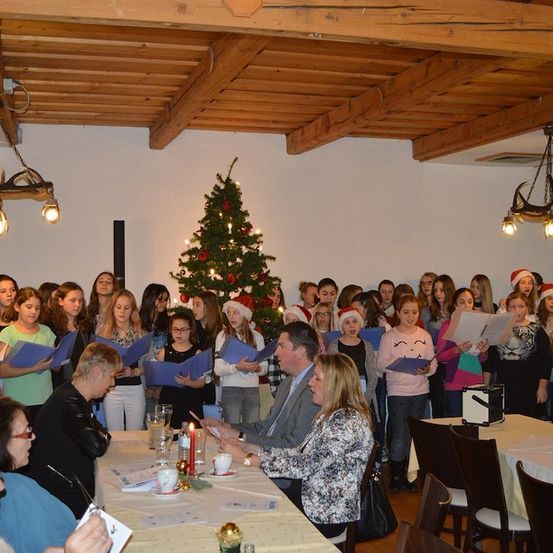 Ein Chor aus Kindern und Erwachsenen in Weihnachtskleidung singt in einem für die Weihnachtszeit geschmückten Raum mit einem Weihnachtsbaum und festlichen Dekorationen.