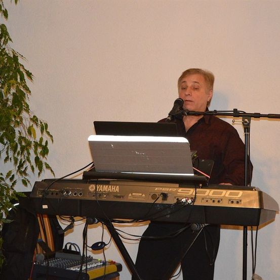 Ein Mann steht vor einem Yamaha-Keyboard und spielt, während er auf einen Laptop schaut, der darauf liegt. Er trägt ein braunes Hemd und schwarze Hose.