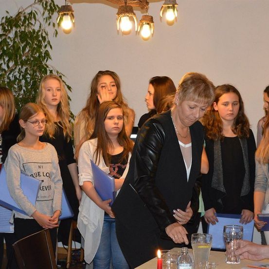 Eine Gruppe von Frauen steht um einen Tisch herum, eine Frau zündet eine Kerze an. Sie halten alle blaue Ordner.