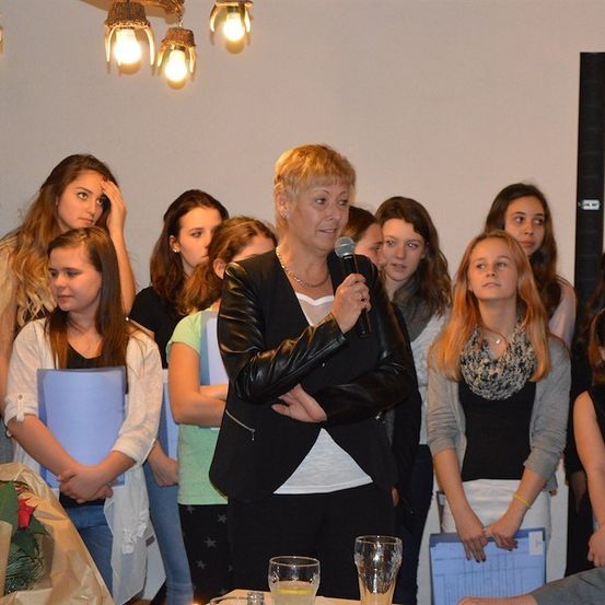 Eine Frau spricht in ein Mikrofon vor einer Gruppe junger Mädchen. Die Mädchen stehen in einer Reihe, und einige halten Ordner. Die Frau trägt eine schwarze Lederjacke und eine Halskette. Im Hintergrund befindet sich eine Pflanze und eine Lampe.