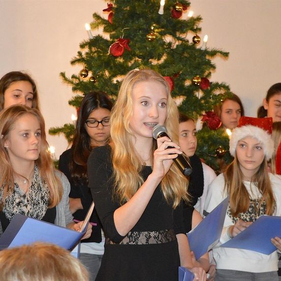 Eine Gruppe junger Mädchen singt Weihnachtslieder vor einem geschmückten Weihnachtsbaum. Das Mädchen in der Mitte hält ein Mikrofon.