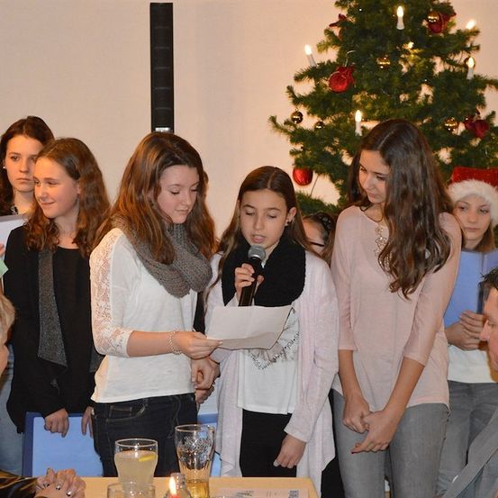 Eine Gruppe junger Mädchen steht um einen Weihnachtsbaum, eine hält ein Mikrofon und liest von einem Stück Papier. Sie sind in Winterkleidung gekleidet, und der Baum ist mit Lichtern und Ornamenten geschmückt.
