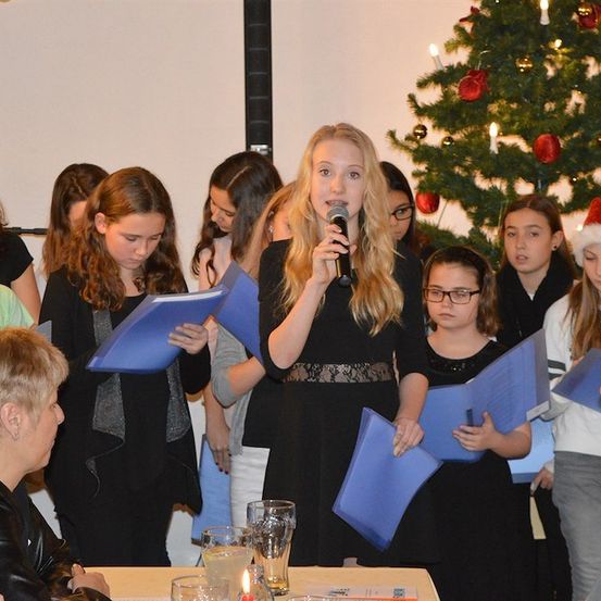 Eine Gruppe von Mädchen und Frauen steht vor einem geschmückten Weihnachtsbaum, hält blaue Papiere in der Hand und ein Mädchen hält ein Mikrofon. Sie singen wahrscheinlich ein Weihnachtslied.