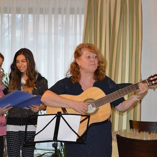 Eine Frau mit roten Locken spielt eine Gitarre, während zwei Mädchen aus einer blauen Mappe lesen. Dahinter sind ein weißer Vorhang und eine grüne Pflanze zu sehen.
