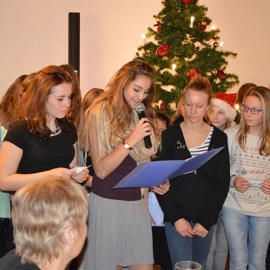 Eine Gruppe junger Mädchen steht um einen Weihnachtsbaum herum, eine hält ein Mikrofon und liest aus einem Skript. Sie sind in Feiertagspullovern und Mützen gekleidet.