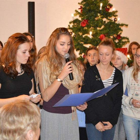 Eine Gruppe junger Frauen steht vor einem Weihnachtsbaum, eine hält ein Mikrofon und liest aus einer blauen Mappe, während andere zuschauen.