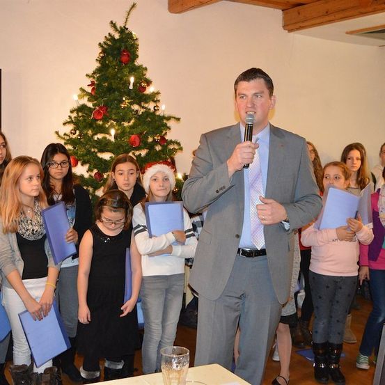 Ein Mann in einem Anzug steht vor einer Gruppe von Kindern mit blauen Ordnern und spricht in ein Mikrofon. Ein geschmückter Weihnachtsbaum steht dahinter.