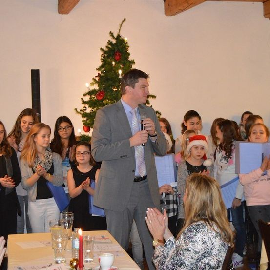 Ein Mann in einem Anzug spricht in ein Mikrofon vor einer Gruppe von Kindern mit Büchern, mit einem geschmückten Weihnachtsbaum im Hintergrund.