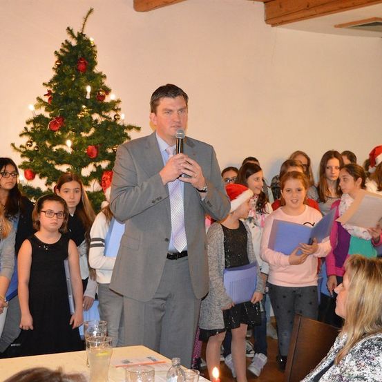 Ein Mann in einem Anzug spricht in ein Mikrofon vor einer Gruppe von Kindern, die Bücher halten und singen, mit einem Weihnachtsbaum im Hintergrund.