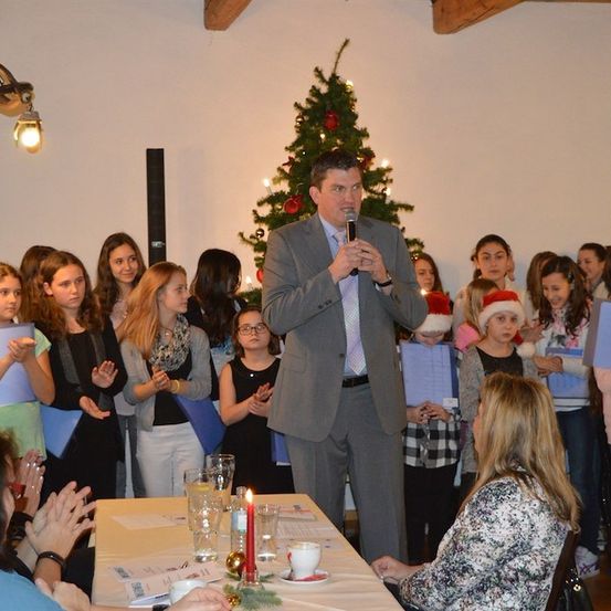 Ein Mann in einem Anzug spricht in ein Mikrofon bei einer Weihnachtsveranstaltung, umgeben von einer Gruppe von Kindern und Erwachsenen, die Bücher halten und klatschen. Ein geschmückter Weihnachtsbaum ist im Hintergrund.
