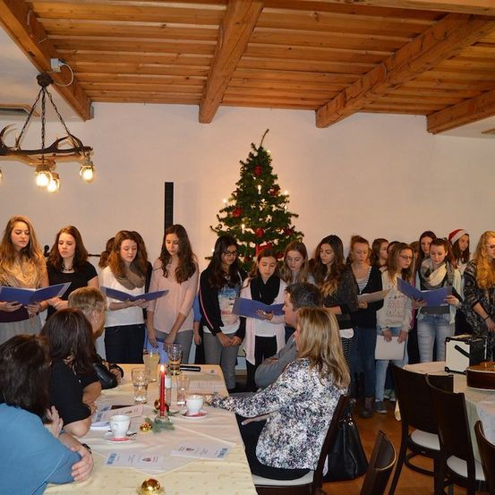 Eine Gruppe junger Frauen steht in einem Raum mit Liederbüchern und singt vor einem Weihnachtsbaum. Eine Frau sitzt an einem Tisch mit einer Kerze und einer Gitarre.